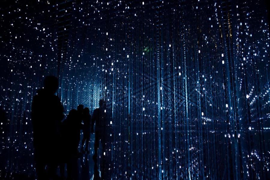 Silhouetted people walk through an immersive art installation of hanging vertical LED light strings in a dark room.